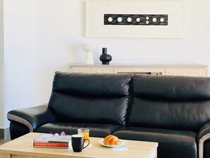 Un canapé en cuir noir dans un salon, avec une table basse en bois sur laquelle sont posés un livre, une tasse et un croissant.
