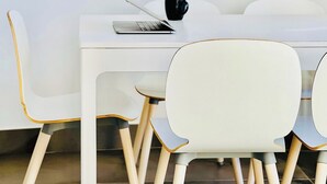 A white dining table with a laptop and a black mug. There are white chairs with wooden legs.