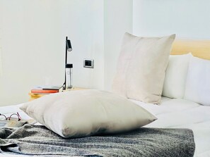 A neatly made bed with a large pillow and a blanket, a bedside lamp, and a small table with books and a glass.