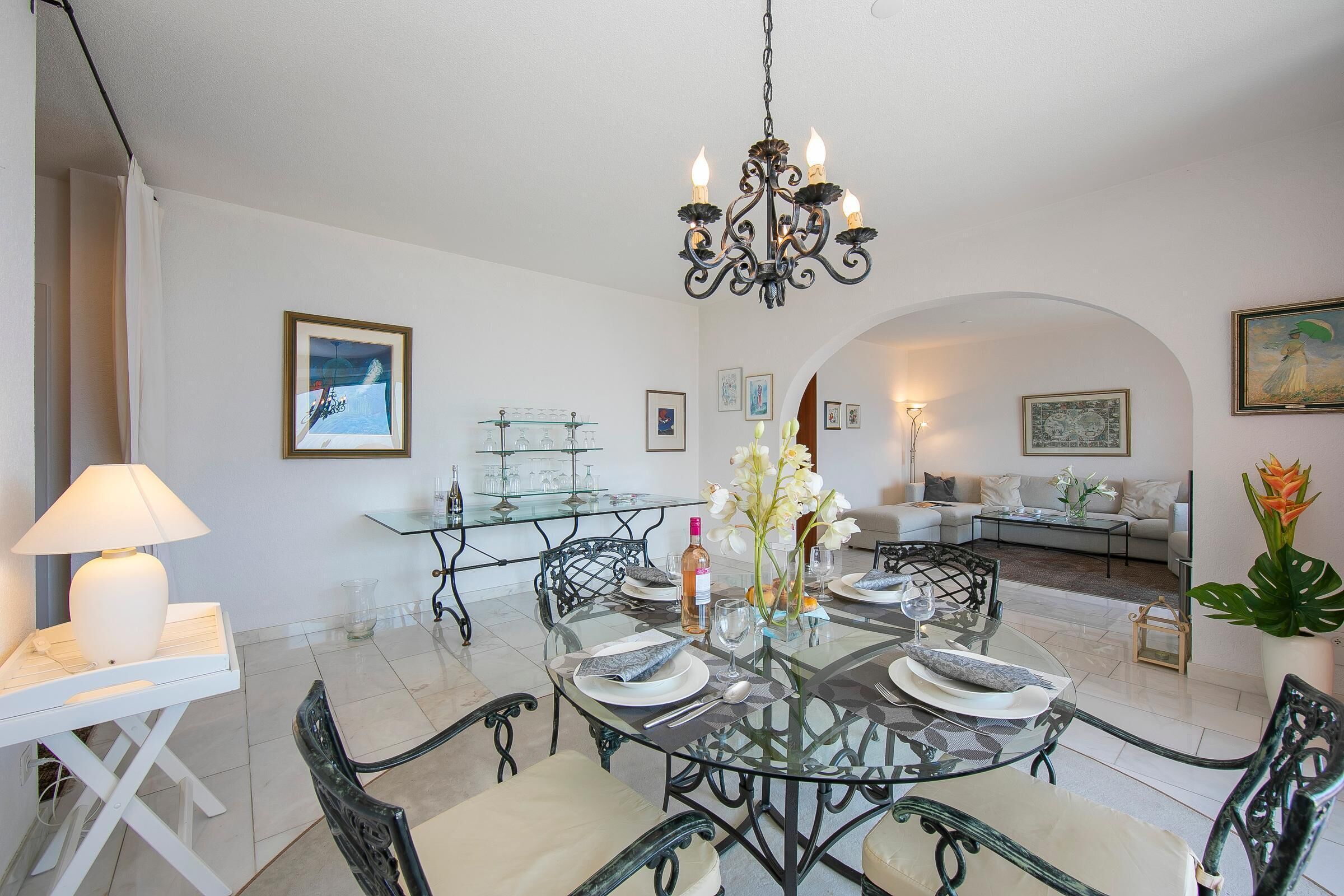 A dining area with a round glass table, black metal chairs, and a chandelier. There is a sofa and a coffee table in the background.