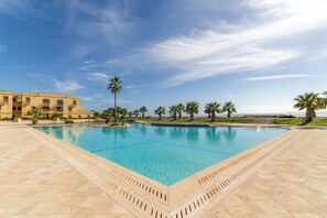 A large rectangular pool with clear blue water, surrounded by a tiled deck and palm trees. - Harbour Views Gozitan Villa Shared Pool (Ghajnsielem)