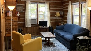 Hirsihirrestä rakennetun mökin sisustus: puulattia, sininen sohva, keltainen nojatuoli, pieni pöytä ja televisio.