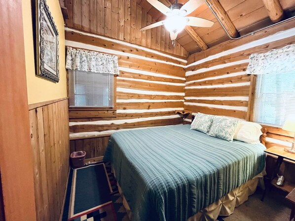 Un dormitorio de cabaña de madera acogedor, con cama, ventana con persianas, ventilador de techo y una mesita.