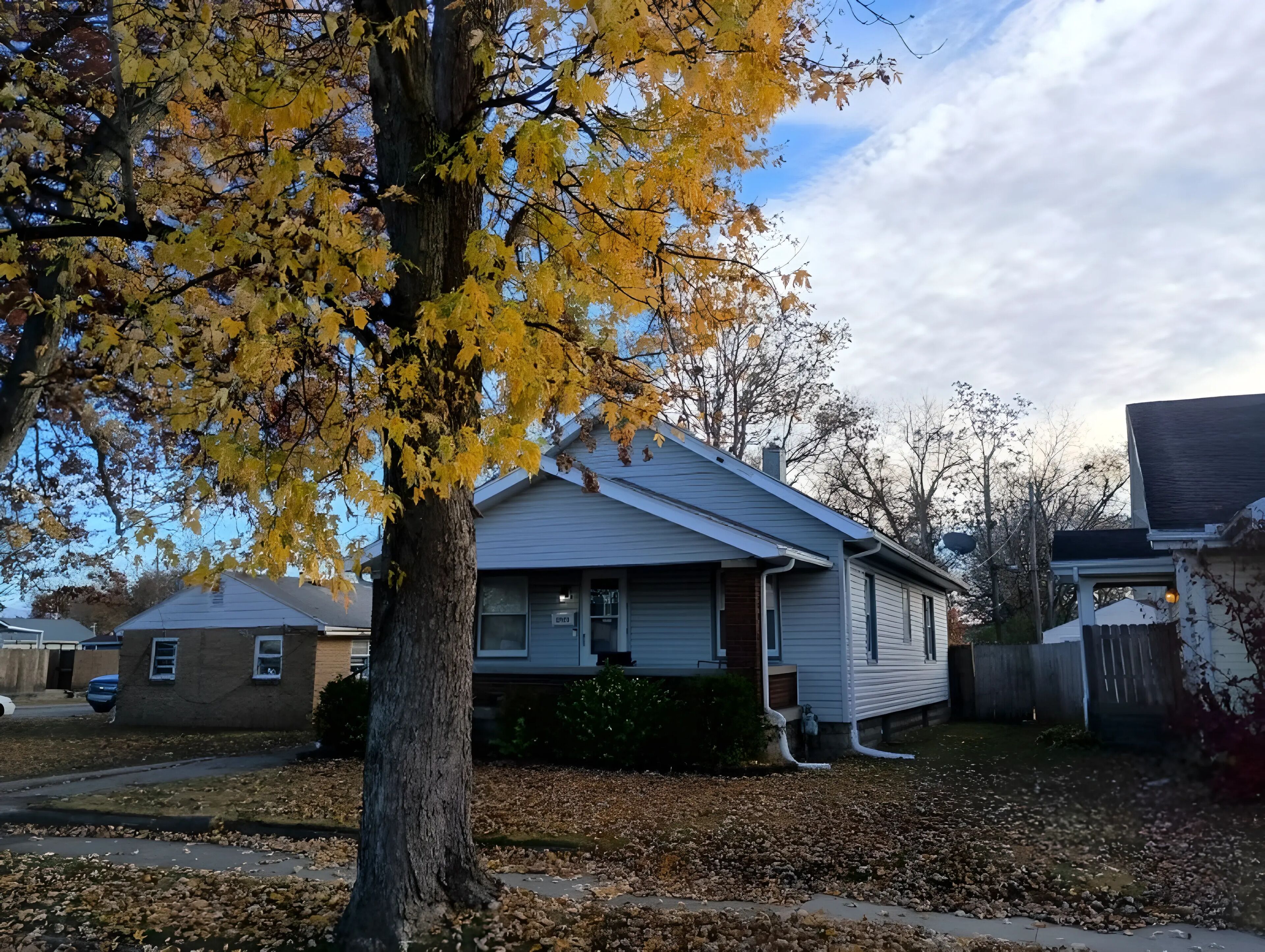 Une maison d’habitation avec un jardin devant, un arbre aux feuilles jaunes et un ciel dégagé.