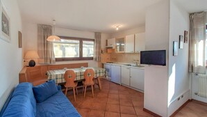 A compact living space with a blue sofa, a dining table with chairs, a kitchen area, and a television. - Nice apartment for 4 guests with TV and pets allowed (Canazei)