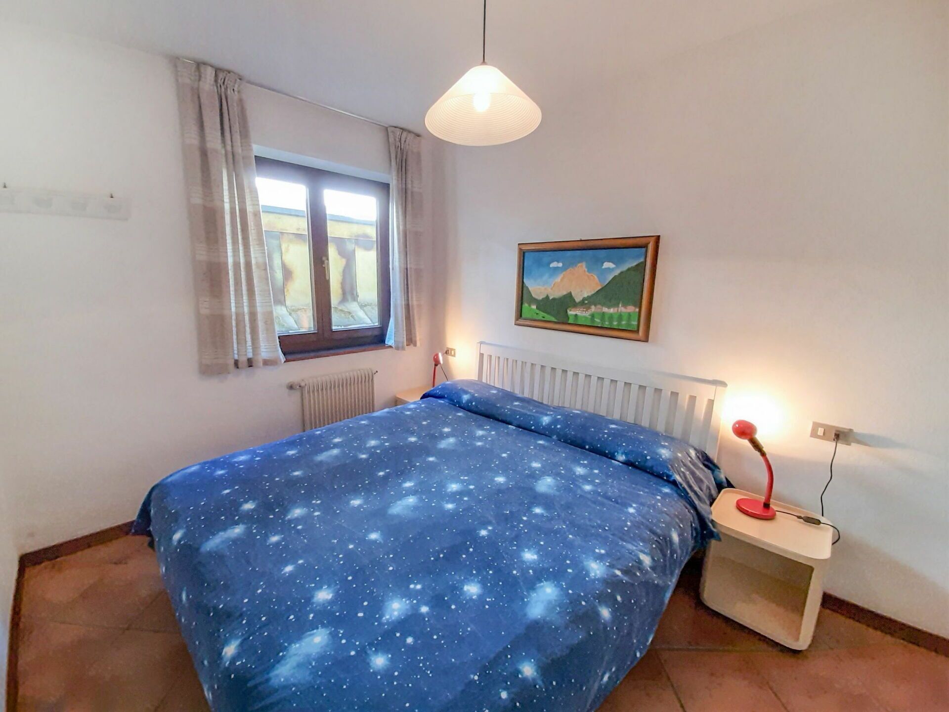 Ein Schlafzimmer mit Bett, Fenster mit Vorhängen, Heizkörper, Nachttisch mit Lampe und einem Bild an der Wand.