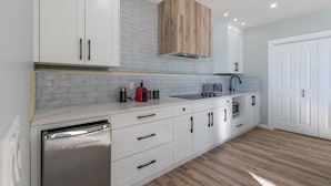 A modern kitchen with white cabinetry, a stainless steel dishwasher, and a wooden upper cabinet. - 1 bedroom and kitchen place with a valley scenery view & free parking (CALGARY)