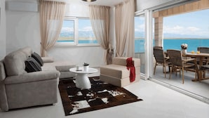 Un salon moderne avec un canapé, une table basse et un coin repas avec des chaises. Une grande fenêtre offre une vue sur la mer.