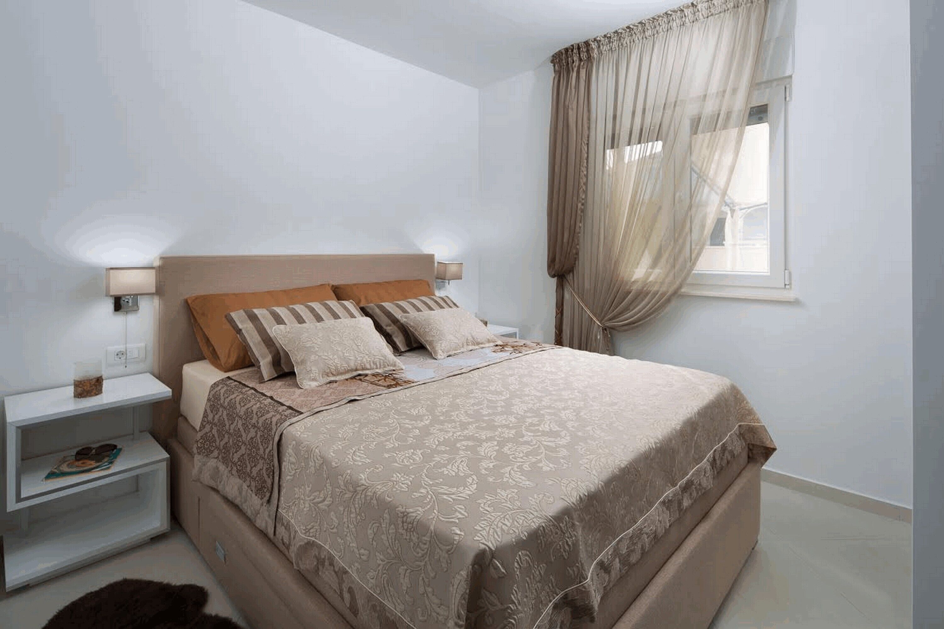 A bedroom with a bed, nightstands, a window with curtains, and a wall-mounted lamp.