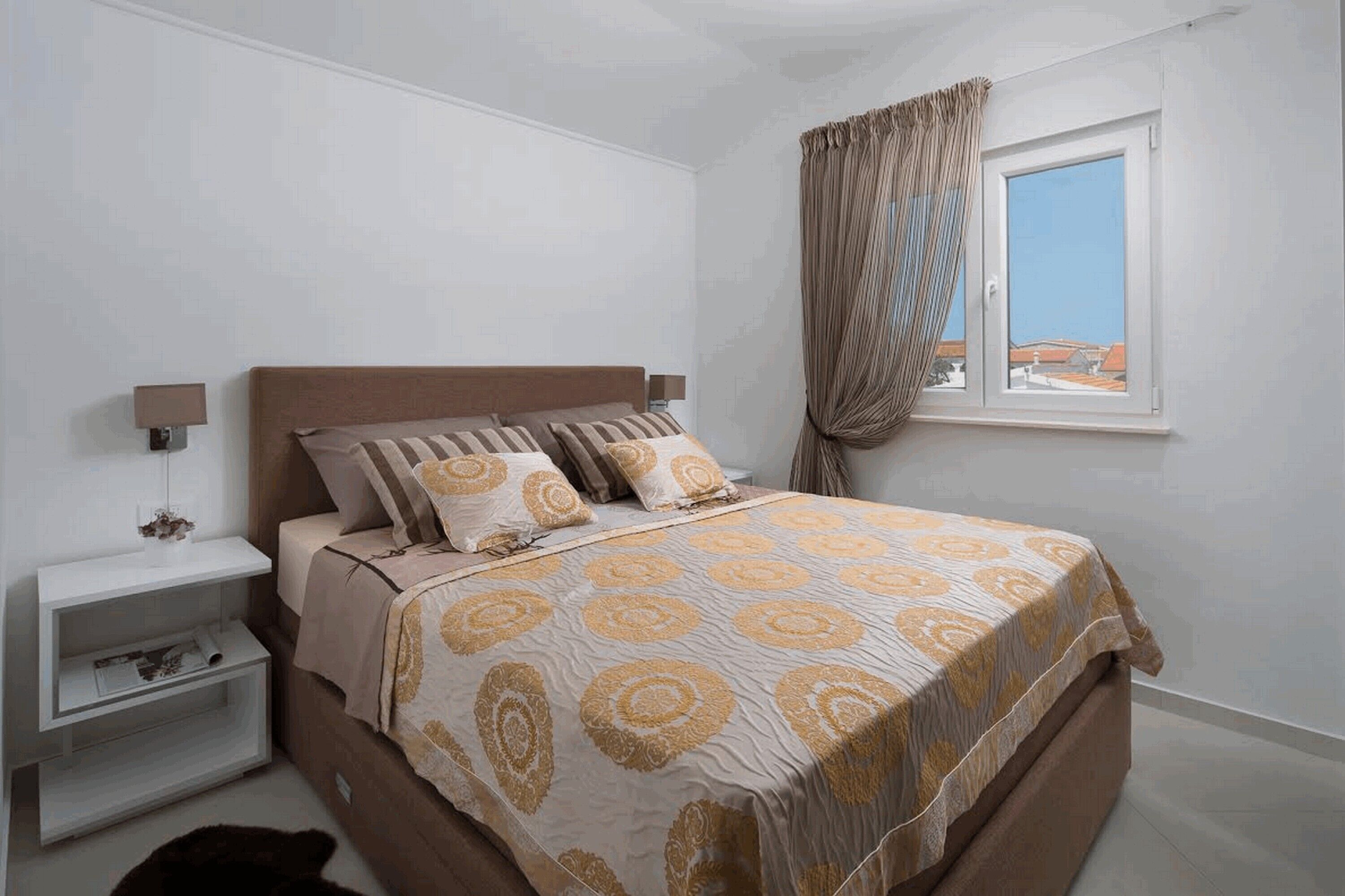A bedroom with a bed, bedside tables, a window with curtains, and a view of buildings outside.