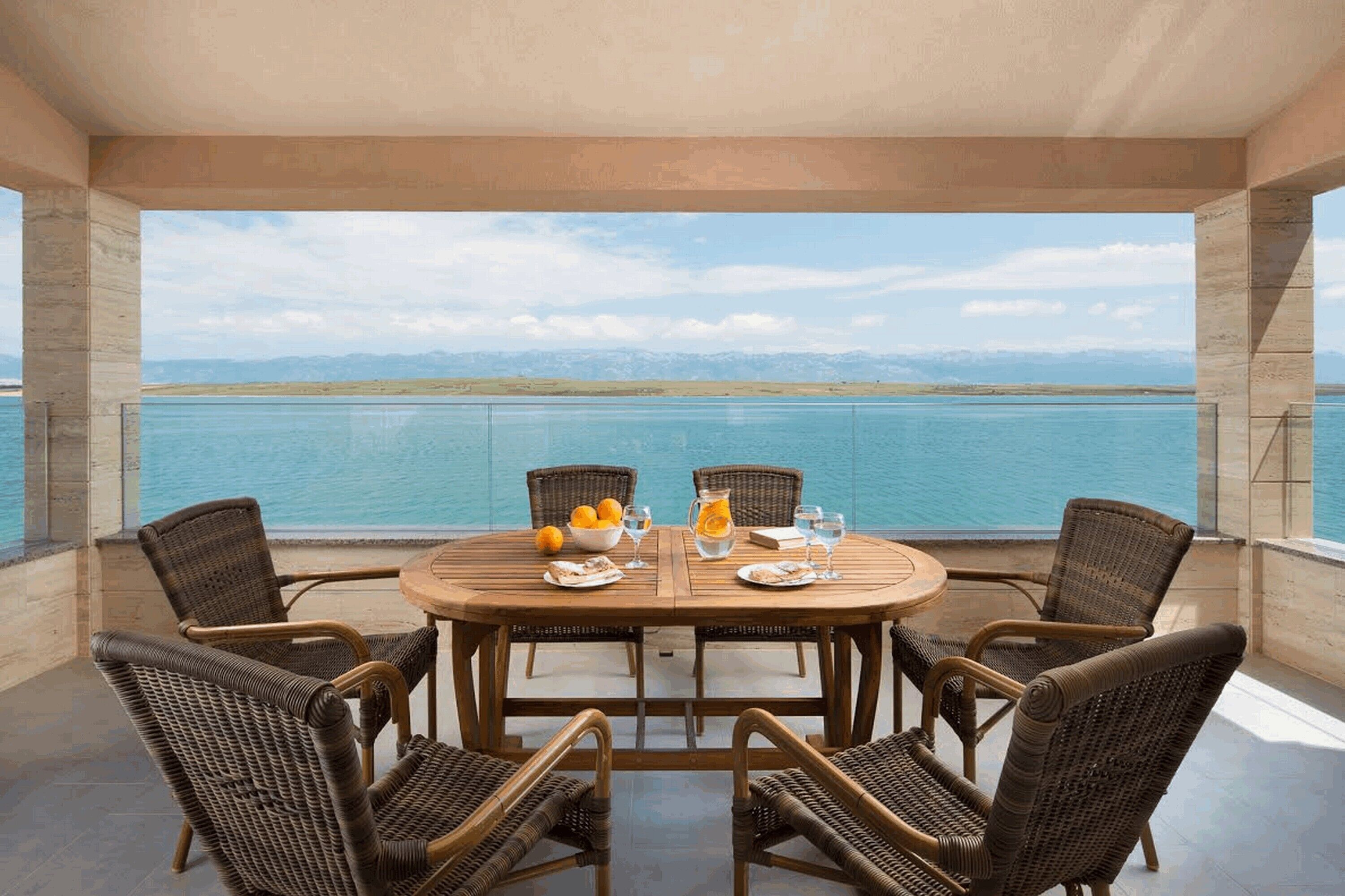A round wooden table with wicker chairs, set against a backdrop of a large body of water and distant mountains.