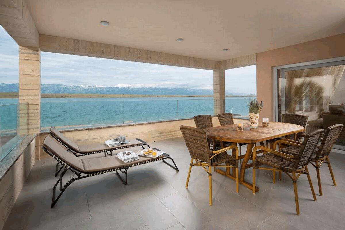 Uma varanda com vista para um lago e montanhas, equipada com uma mesa, cadeiras e espreguiçadeira.