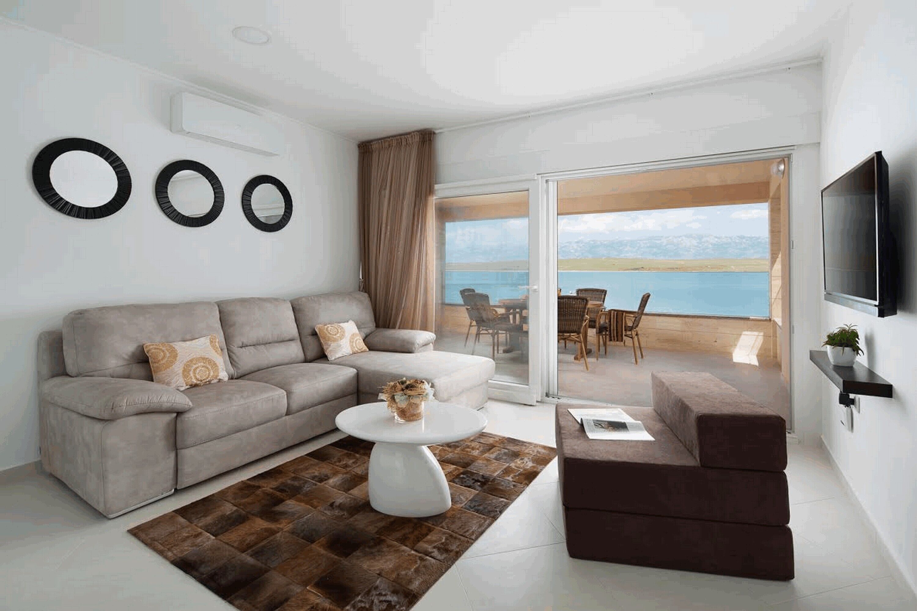 A modern living room with a grey sofa, a round coffee table, and a brown ottoman. There is a TV mounted on the wall and a large window offering a view of the sea.