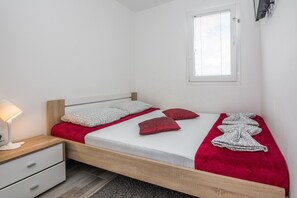 Un dormitorio con cama de madera, ropa de cama blanca, almohadas rojas, una mesita de noche con lámpara y una ventana con persianas.