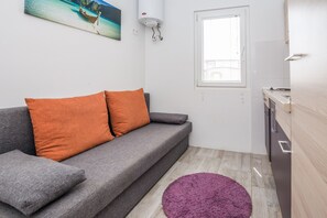 Un espacio habitable pequeño con un sofá gris, dos cojines naranjas, una alfombra morada, una pequeña zona de cocina y una ventana con persianas.