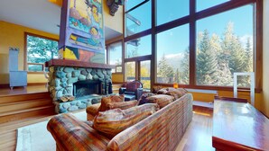 A cozy living room with a stone fireplace, large windows, and a view of trees. - Stunning 3 Bedroom / 3.5 Bathroom Blueberry house with breathtaking Mountain Views (Whistler)