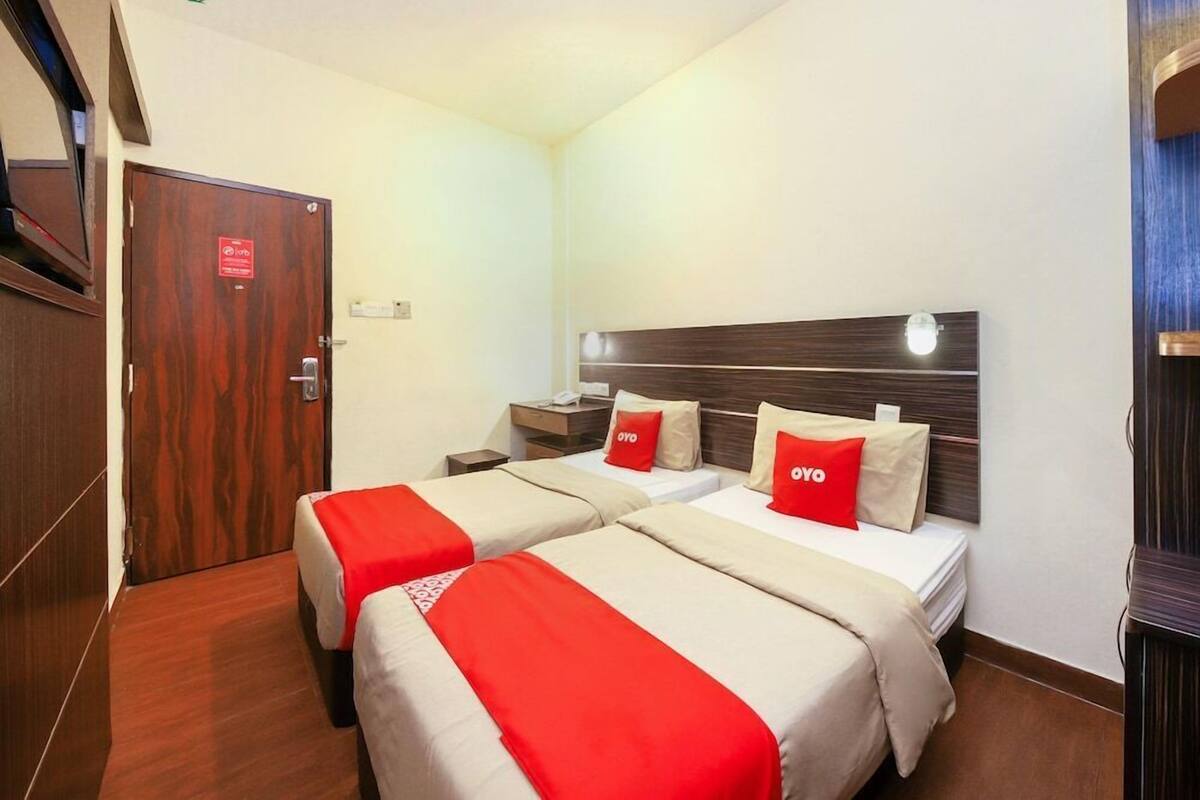 A hotel room with two beds, a wooden door, a nightstand, and a wall-mounted lamp.