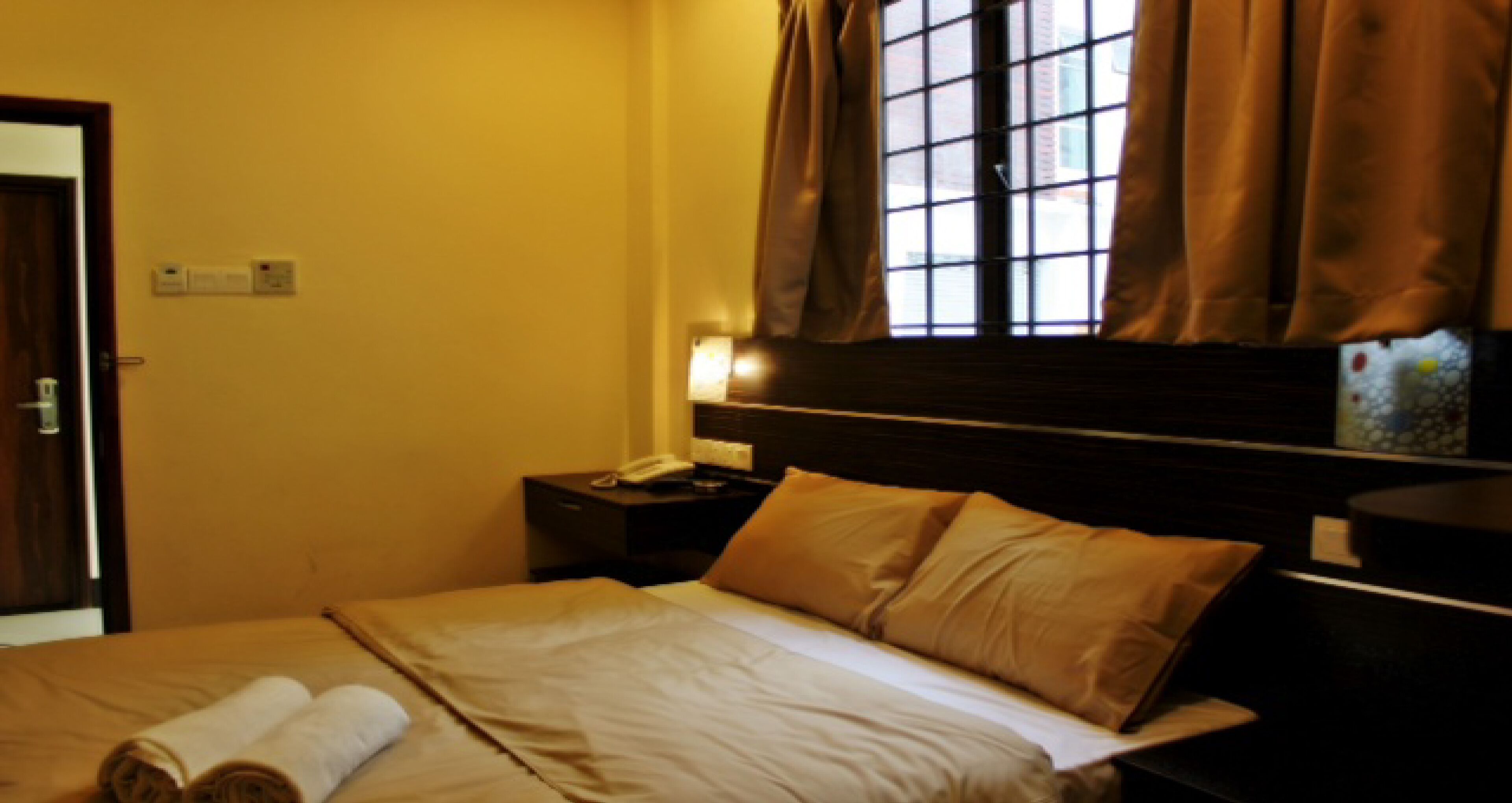 Quarto de hotel com uma cama, mesa de cabeceira com abajur, janela com cortinas e uma porta.