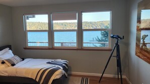 A bedroom with a bed, a nightstand, a lamp, a tripod, and a window with a view of a lake and forest. - Waterfront Home w/ Mt Rainier Views, Private Beach, 4 Decks, Sleeps 9 (Bremerton)
