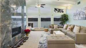 A spacious living room with a large window overlooking a body of water, a fireplace, and a ceiling fan. - Waterfront Home w/ Mt Rainier Views, Private Beach, 4 Decks, Sleeps 9 (Bremerton)