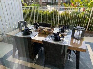 Un área de comedor al aire libre con una mesa de vidrio, sillas de mimbre y una valla metálica al fondo.