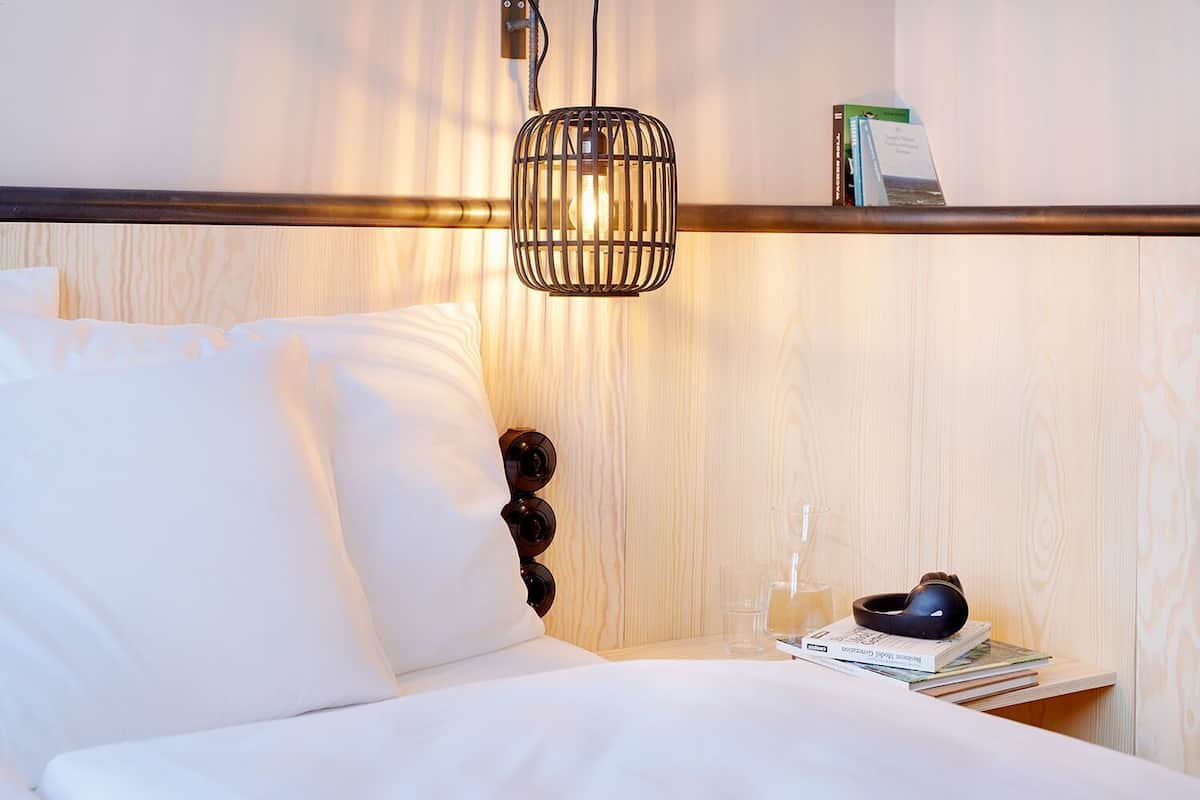 Una habitación de hotel con cabecera de madera, una mesita de noche con lámpara, vaso y libros.