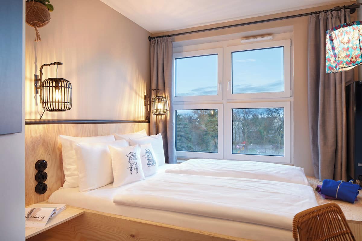 Habitación de hotel con una cama grande, una mesita de noche con lámpara y vistas al exterior a través de una ventana.