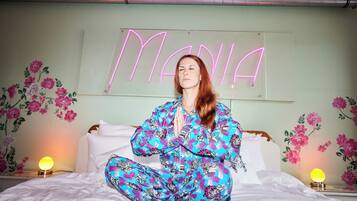 A person sitting on a bed in a room with floral wallpaper and a neon sign that reads "MANIA".