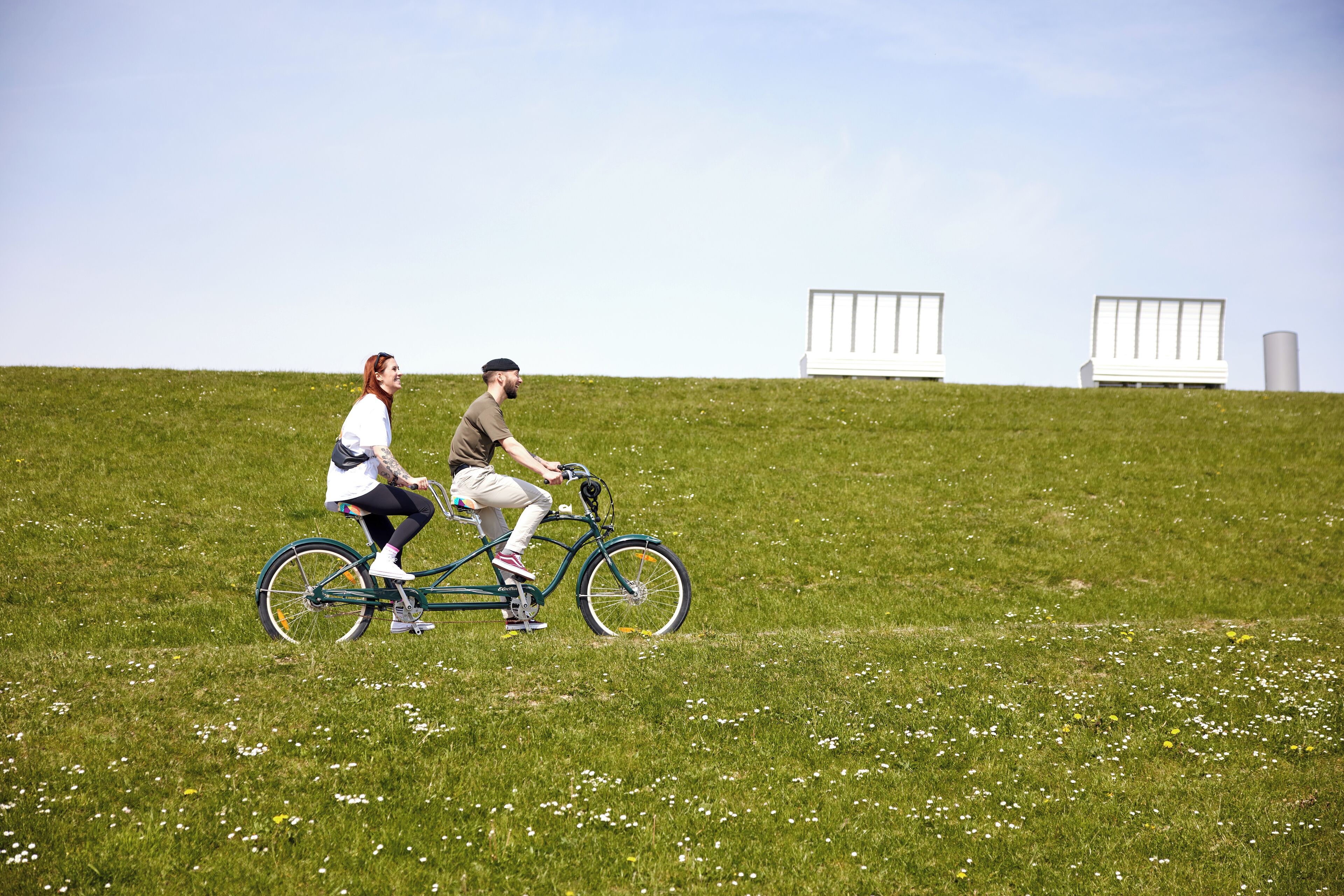 Dos personas andando en bicicleta sobre un campo césped con estructuras blancas al fondo.