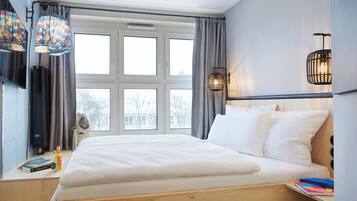 Ein Hotelzimmer mit einem großen Bett, einem Fenster mit Vorhängen, einem Nachttisch mit Büchern und einer Flasche sowie zwei Wandlampen.
