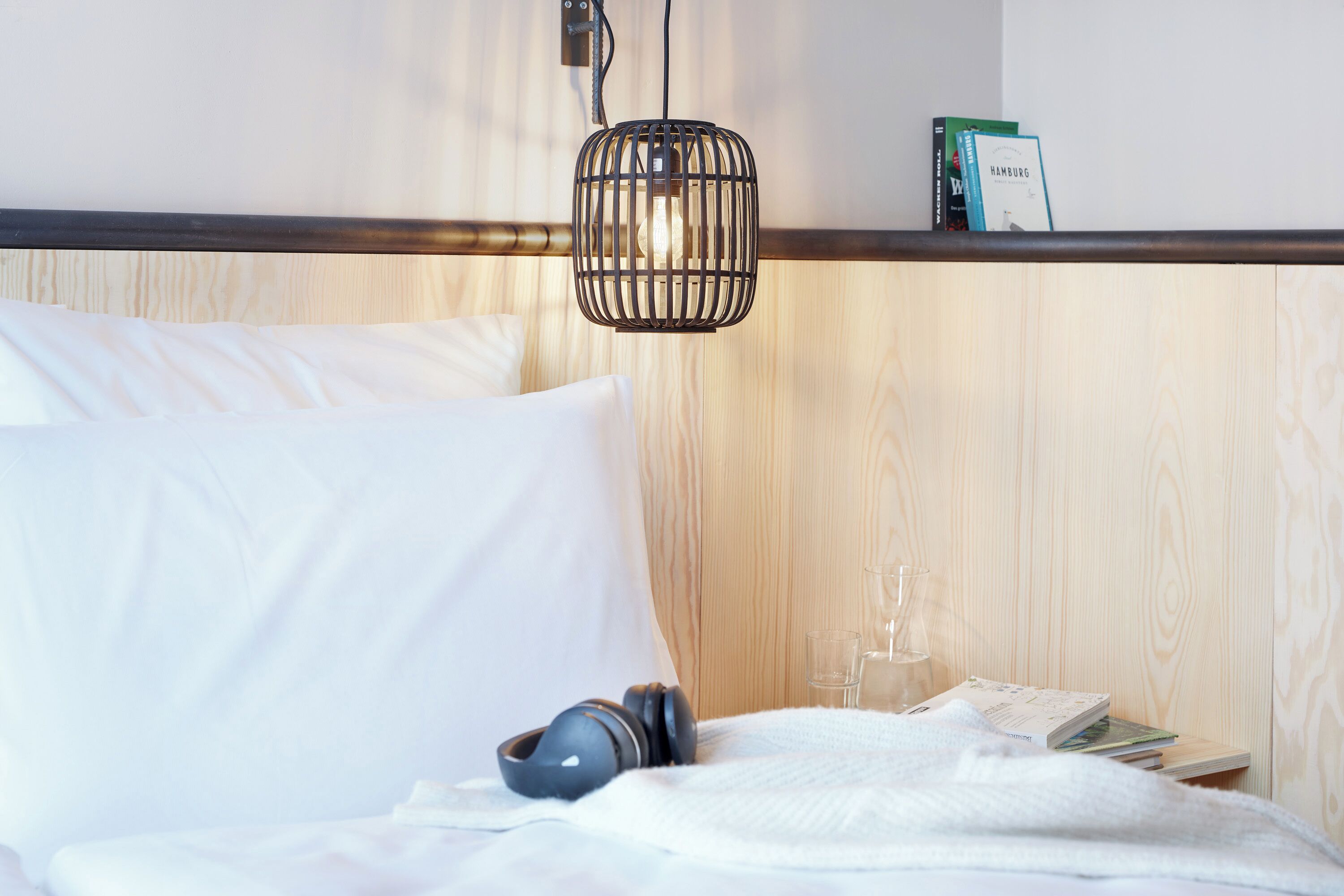 A neatly made bed with white linens, a pair of headphones, a glass of water, and a book on a bedside table.