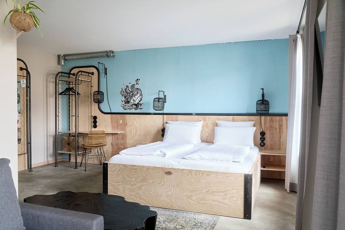 Un dormitorio moderno con cabecera de madera, cama con ropa blanca, una pequeña mesa con una silla y un estante en la pared.