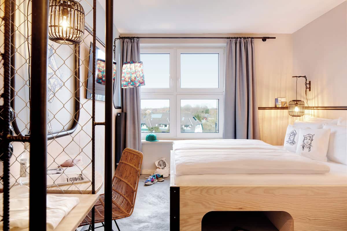Habitación de hotel con cama, una silla, ventana con cortinas e iluminación en la pared.