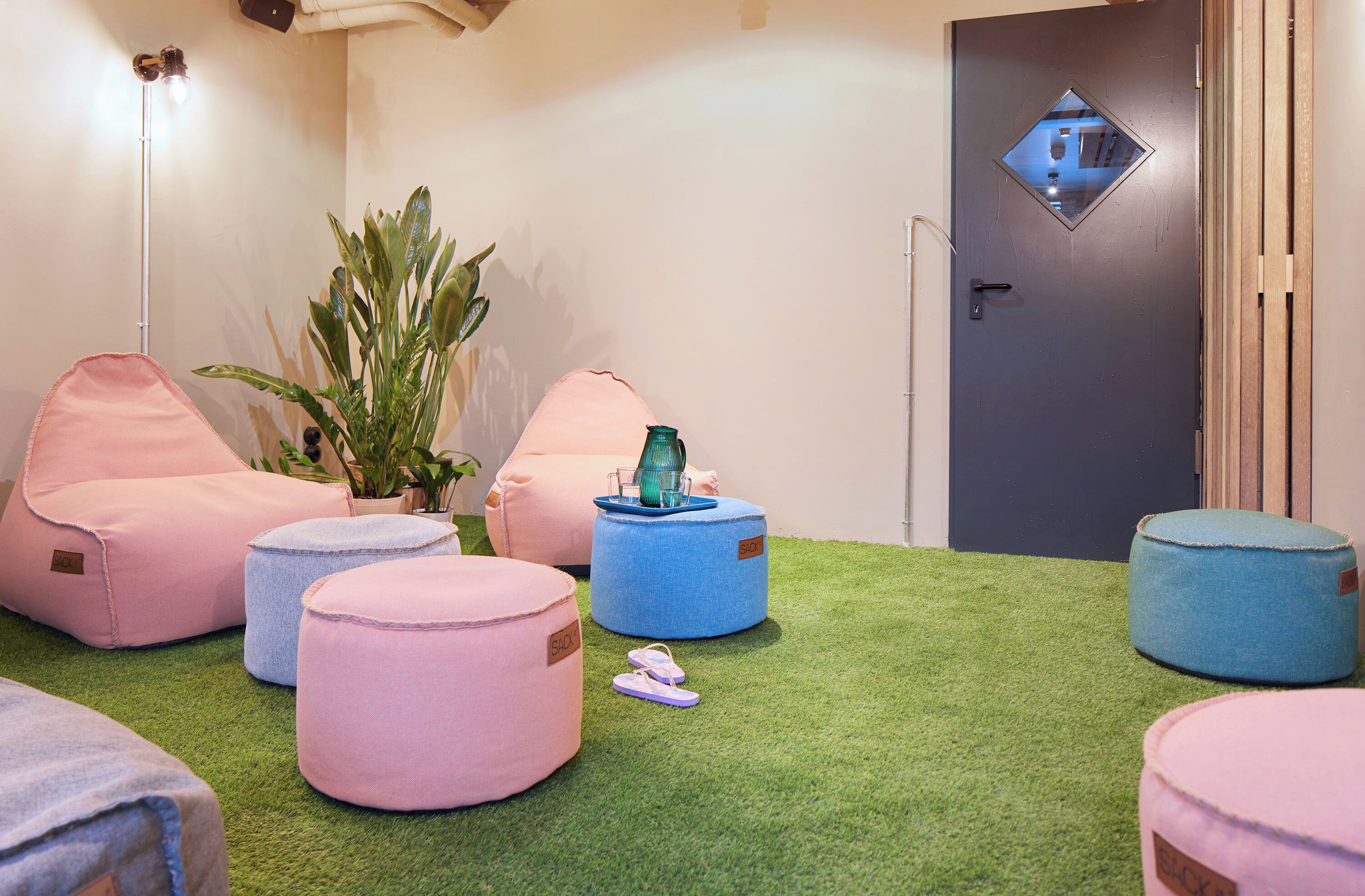 A room with green artificial grass, pink and blue bean bags, a potted plant, and a door with a diamond-shaped mirror.