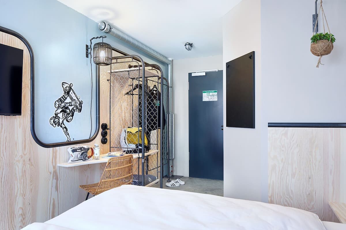 Una habitación de hotel moderna con cama, escritorio con silla, planta colgante, armario y puerta con un letrero.