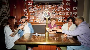 Eine Gruppe von Personen sitzt an einem Tisch, in dessen Mitte sich eine Uhr befindet. Die Wand um sie herum ist mit Fotos geschmückt.