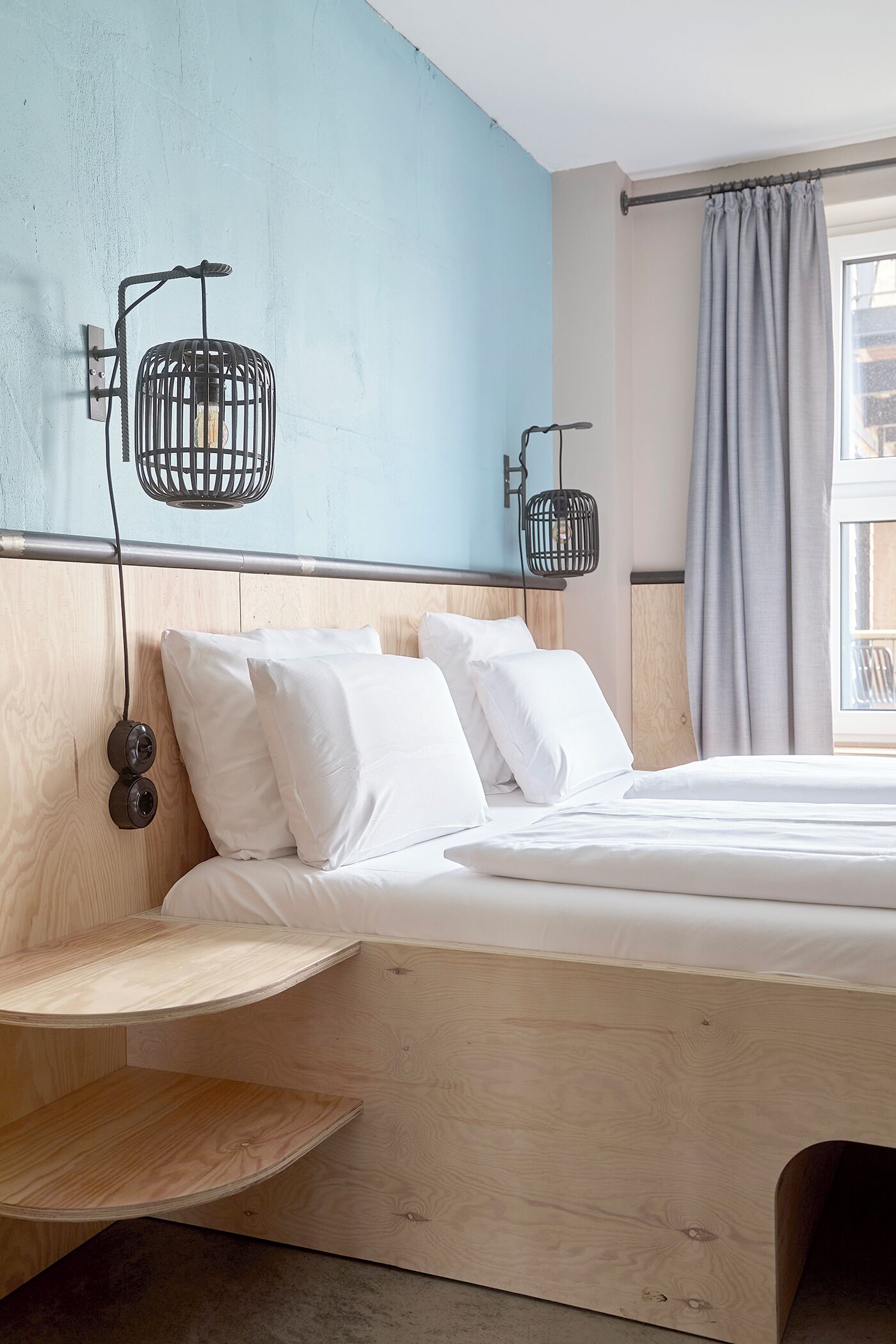 Un dormitorio moderno con cabecera de madera, cama con ropa de cama blanca, dos lámparas fijadas a la pared y una ventana con cortina.