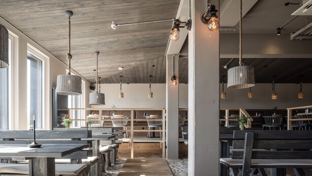 A spacious indoor area with wooden benches and tables, hanging wicker lamps, and a visible staircase leading to another level. - Bretterbude Büsum (Büsum)