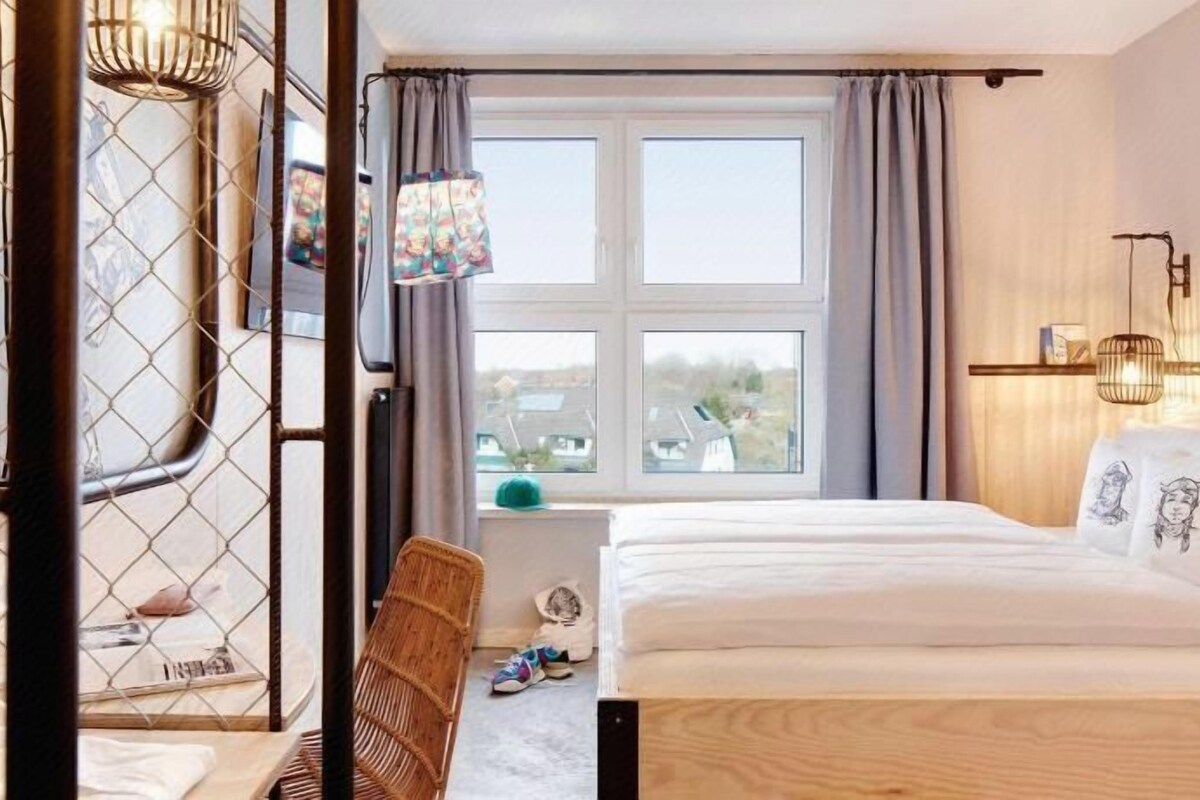 A hotel room with a bed, a chair, a window, and a wall-mounted light fixture.