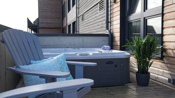 A hot tub with a control panel, a lounge chair with patterned cushions, and a potted plant on a wooden deck.