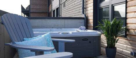 A hot tub with a control panel, a lounge chair with patterned cushions, and a potted plant on a wooden deck.