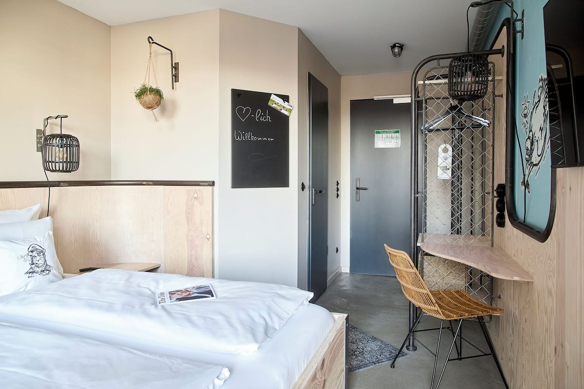 Habitación de hotel con cama, una mesita, una silla, una planta colgante y un cartel de bienvenida.