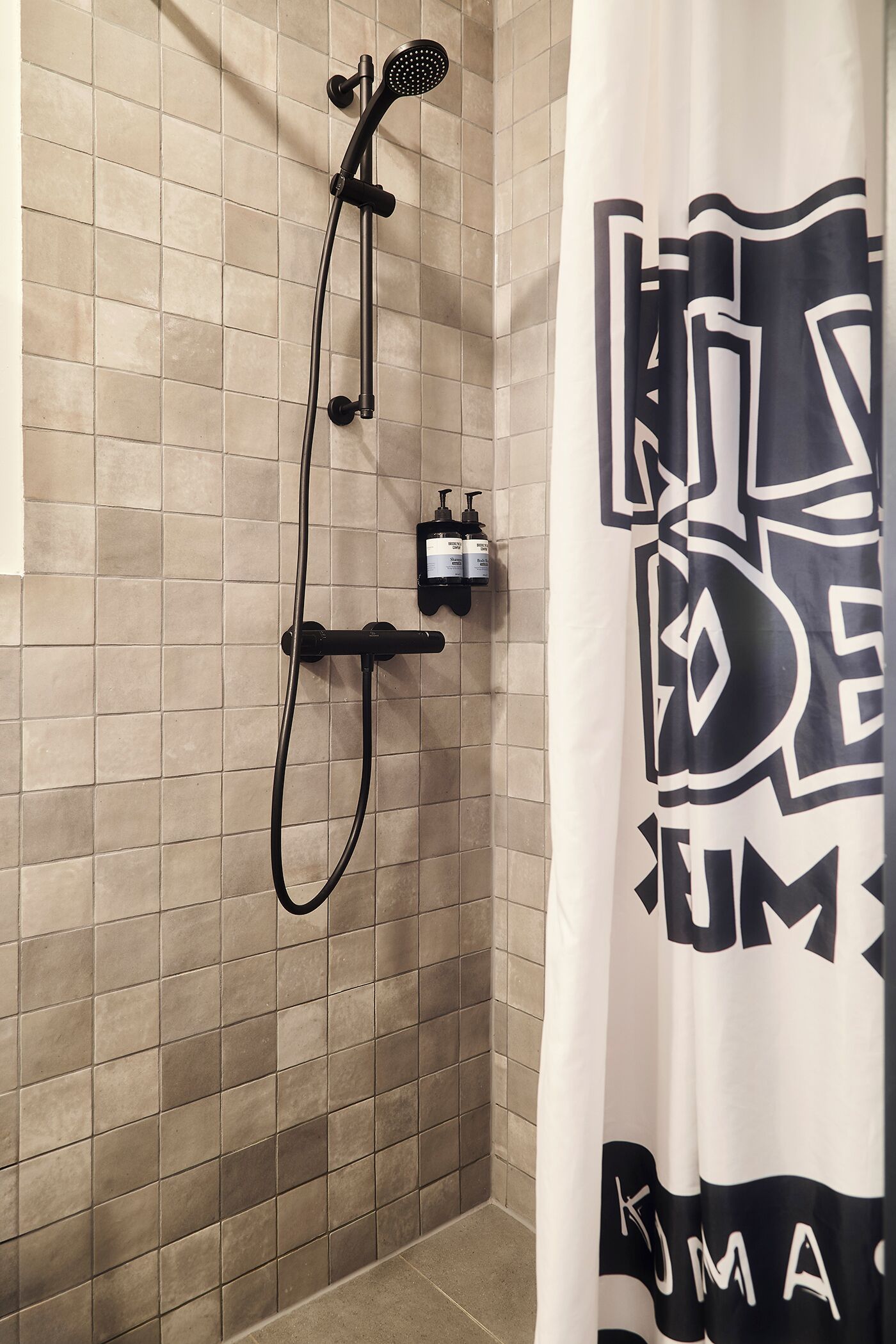 Zona de ducha con cabezal de ducha de mano, panel de control en la pared y cortina de ducha con estampado blanco y negro.