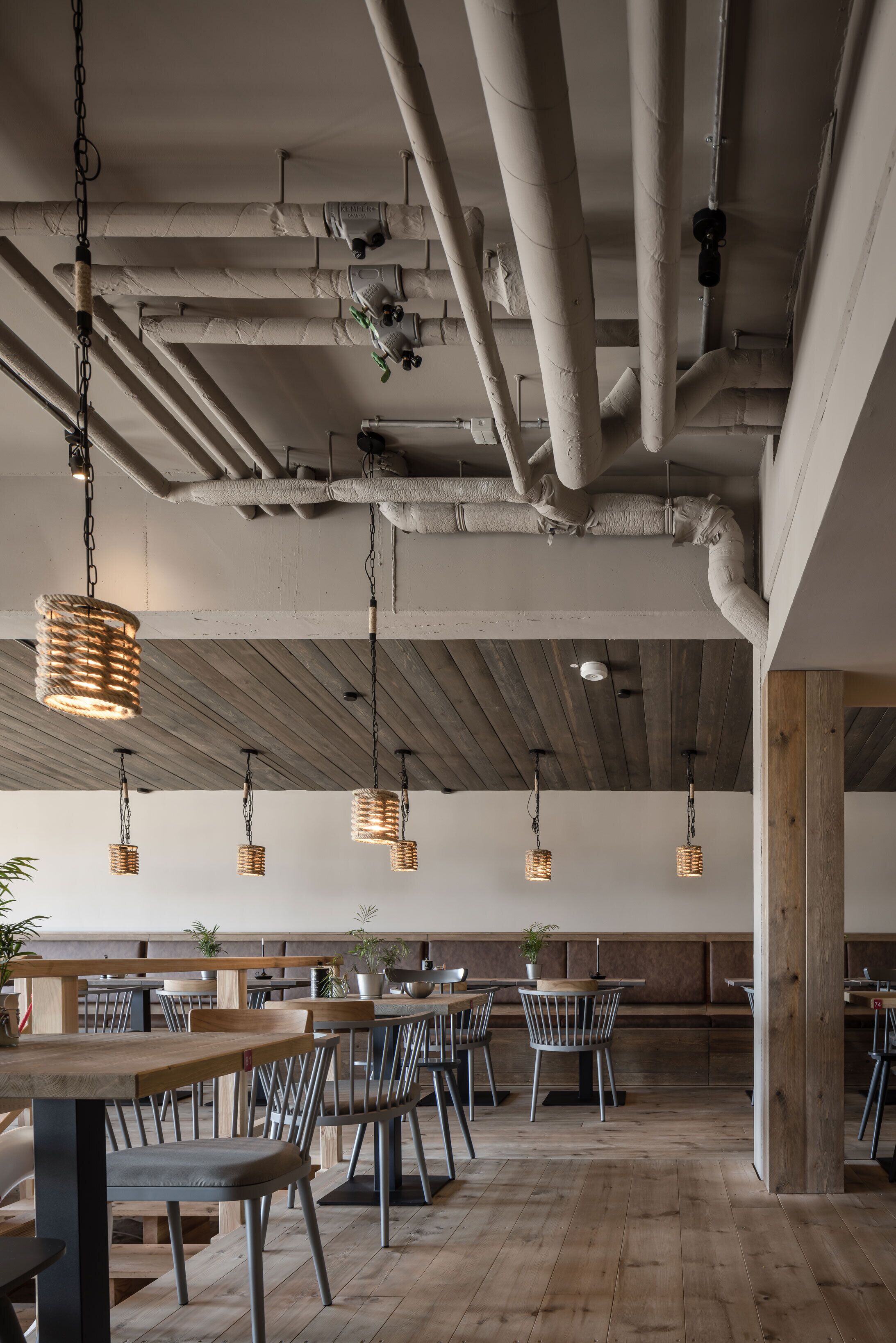 Un grand espace dédié à la salle à manger, avec des tables et des chaises en bois, des tuyaux apparents et des lampes suspendues en osier.