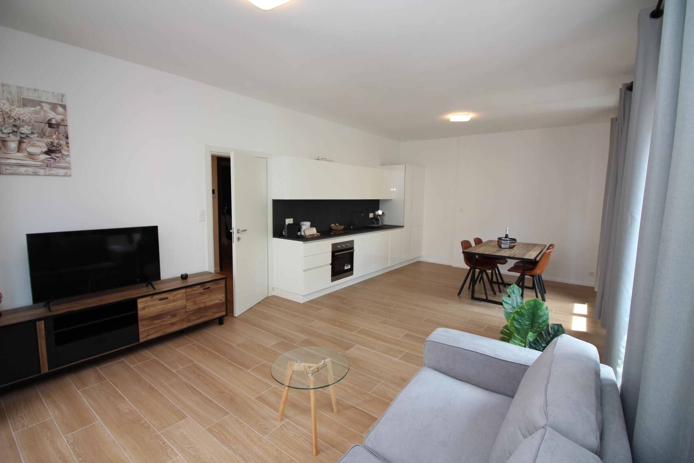 A modern living room with a flat-screen TV, a grey sofa, a glass coffee table, and a dining area with a wooden table and chairs.