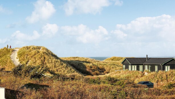 Paesaggio costiero con dune, una casa dal tetto scuro e rivestimenti verdi, un sentiero sterrato che sale sulla duna e un'auto parcheggiata nelle vicinanze.