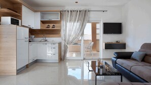 Una cocina moderna con armarios blancos, un microondas y un lavamanos. Hay una zona de comedor con una mesa y sillas negras, y un sofá con un televisor montado en la pared.