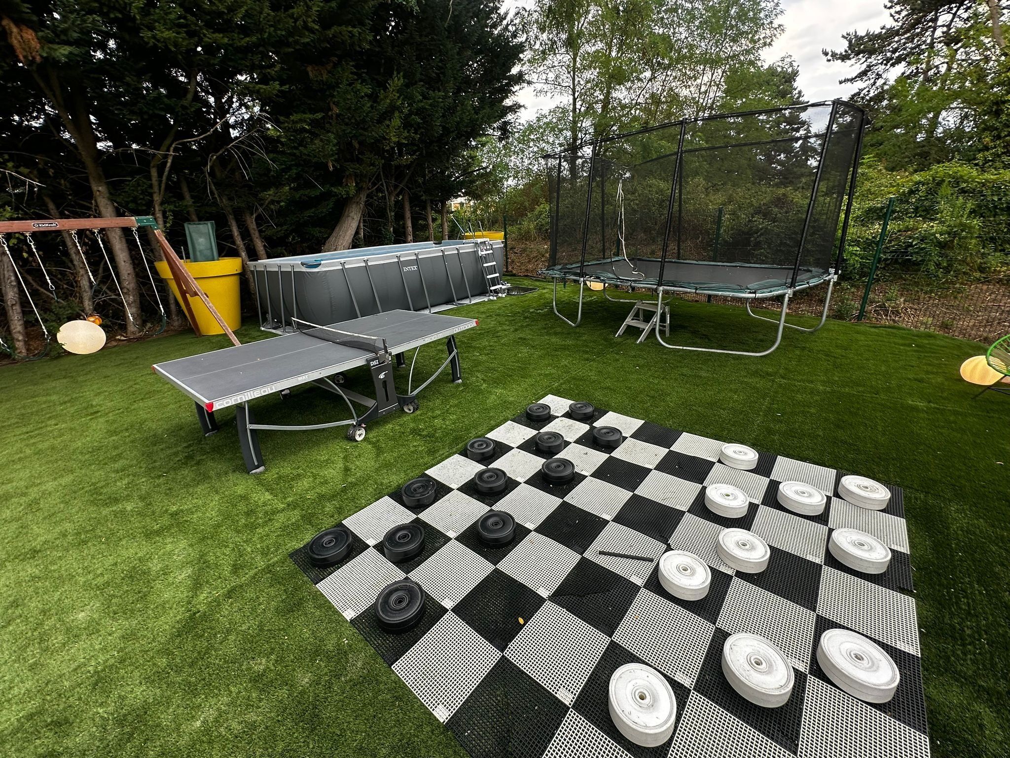 A backyard with a ping pong table, a checkered table tennis table, and a trampoline.