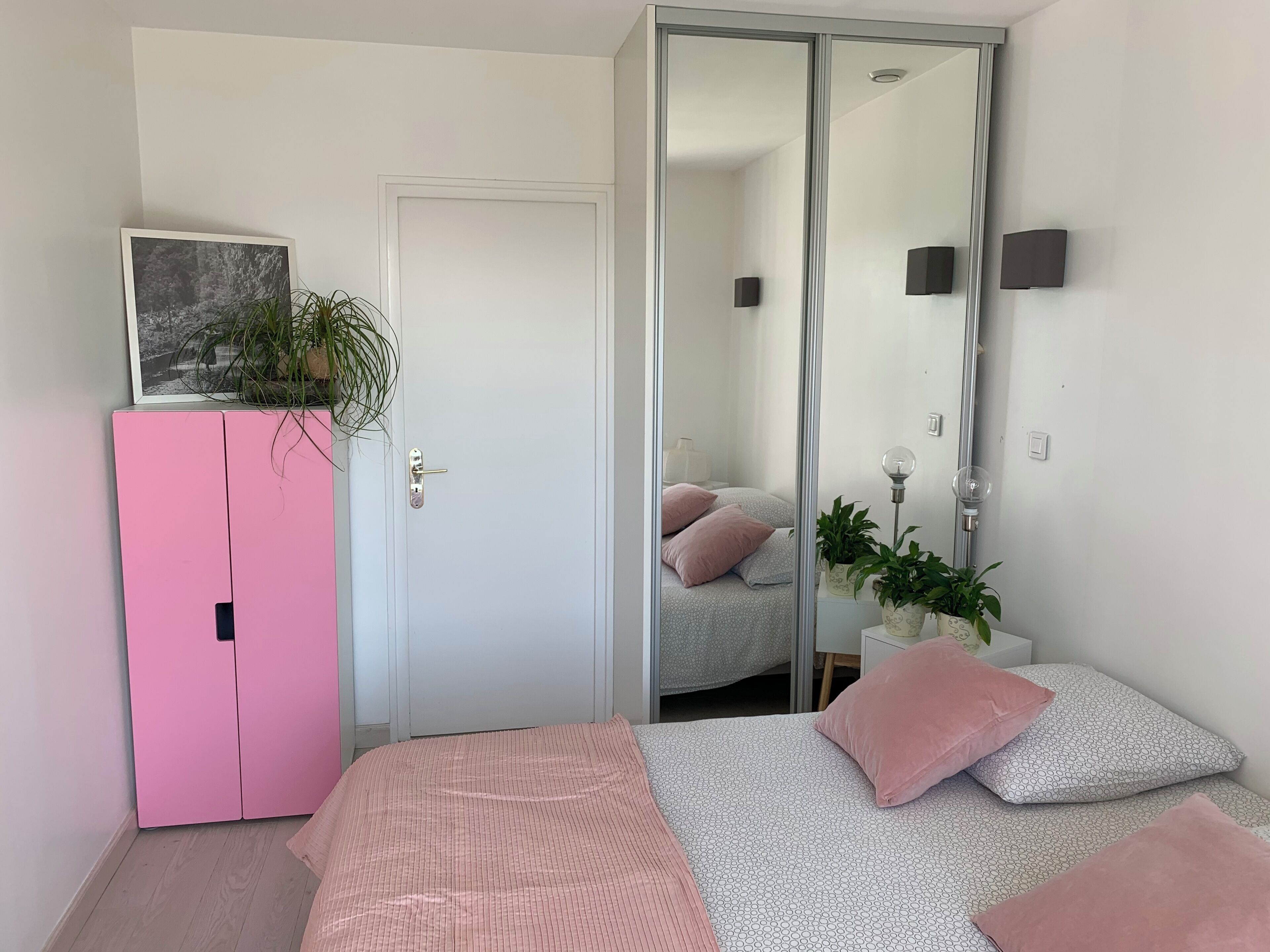 A bedroom with a bed, a pink wardrobe, a sliding door, a mirror, and a potted plant.