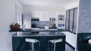 A modern kitchen with white cabinets, a black countertop, a built-in microwave, and a coffee machine. There are two bar stools and a potted plant on the counter. - Casabina (Chelles)
